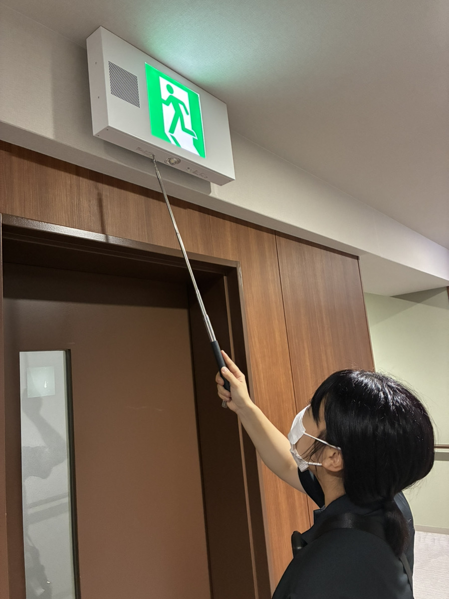 女性消防設備士の草地が点検棒で点滅形誘導灯を点検する様子