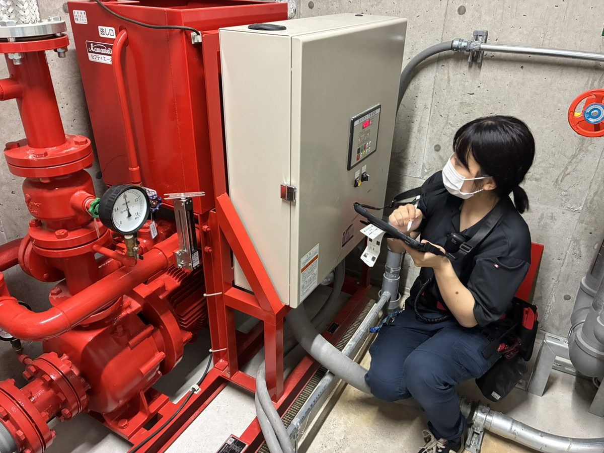 消火ポンプ室で消火ポンプの制御盤をiPadで確認する女性消防設備士の草地